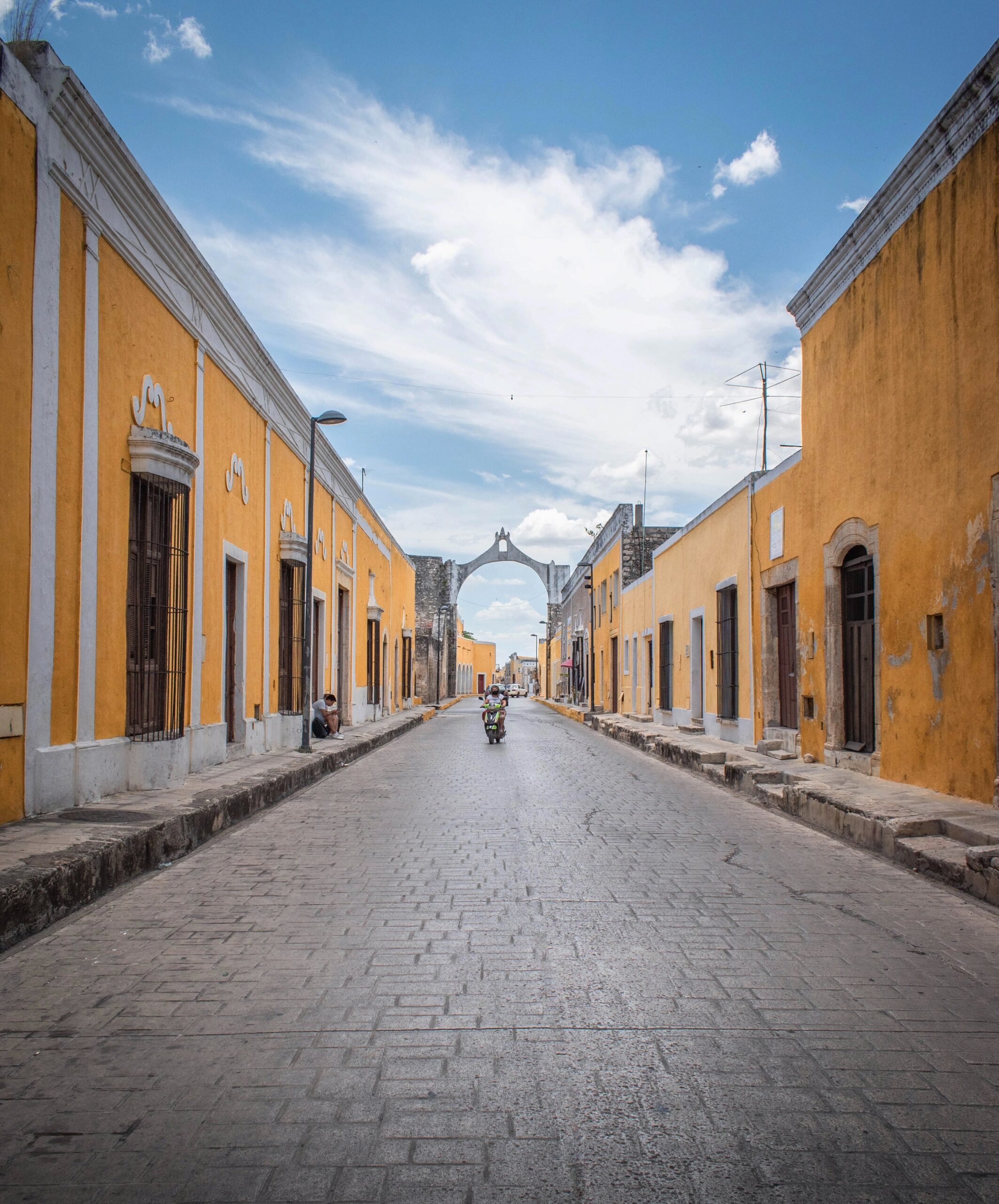 A street of izamal with yellow buildings on sides and a woman on a scooter in the center